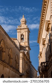A Baroque bell tower rises between limestone facades in Mdina, Malta. Midday light sharpens arched windows, carved cornices, and honey stone with crisp shadows.
