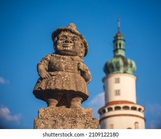 Baroque Stony Figure Of The Dwarf Cabinet Near Castle Of Nove Mesto Nad Metuji, Czech Republic. Castle Tower On Background. Dwarf Cabinet Is Mini Sculptures Created Baroque Sculptor In 18th Century