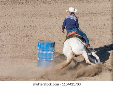 Barrel Racing A cowgirl with long blond braid, white hat and blue shirt rides on the back of a white horse turns her horse around the backside of a blue barrel in a barrel racing contest at a rodeo.