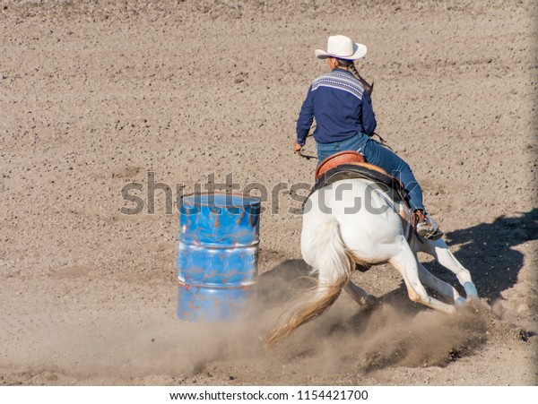 Barrel Racing A cowgirl with long blond braid,\
white hat and blue shirt rides on the back of a white horse turns\
her horse around the backside of a blue barrel in a barrel racing\
contest at a rodeo.