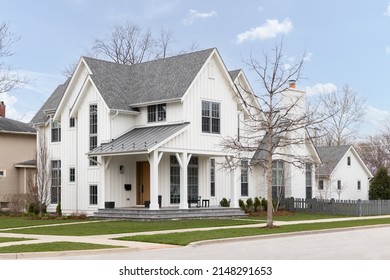 BARRINGTON, IL, USA - APRIL 20, 2022: A White Modern Farmhouse With A Covered Front Porch, Board And Batten Siding, A Wooden Front Door, And Grey Fence.