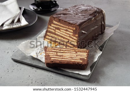 Bar-shaped chocolate cake with biscuits layered inside and chocolate icing. Uncooked dessert Kalter Hund or Hedgehog Slice. Served on white paper on grey tray. Viewed in close-up on grey table surface