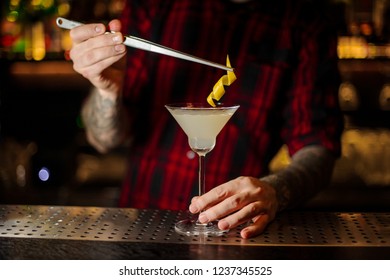 Bartender adding to a White Lady cocktail in the glass one orange zest with tweezers on the bar counter