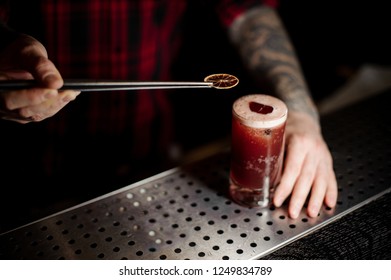 Bartender Decorating A Sippy Cup Cocktail In The Glass With One Dried Orange With Tweezers On The Bar Counter