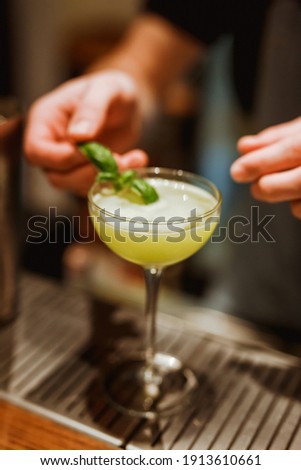 Bartender garnishing a fresh sour cocktail in a coupe glass with a basil leaf. Lifestyle vertical image. Selective focus.