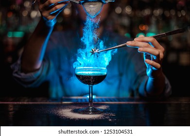 Garota bartender adicionando a um coquetel marrom e despeje em um badian flamejado em pinças um açúcar em pó no balcão da barra na luz azul.