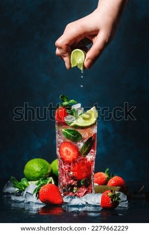 Bartender hand squeezes lime juice into strawberries mojito cocktail glass with lime, white rum, soda, cane sugar, mint, and ice in glass on deep blue background. Summer refreshing beverage  商業照片 © 