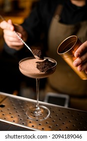 Bartender Holding Metallic Jar And Spoon In His Hands And Decorating Brown Cocktail Standing On Metal Bar Counter With Chocolate