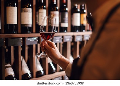 Bartender or male cavist standing near the shelves of wine bottles holds a glass of wine, looks at tint and smells flavor of wine in glass.