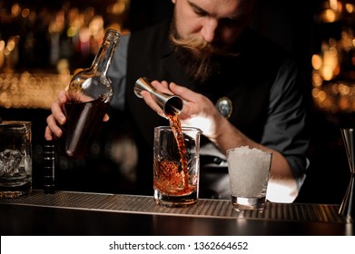 Bartender pouring a brown alcohol to the cocktail from the steel jigger to a glass on the bar counter on the blurred background