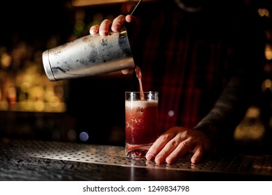 Bartender Pouring A Delicious Sippy Cup Cocktail From The Steel Shaker To A Glass With Big Ice Cube On The Bar Counter