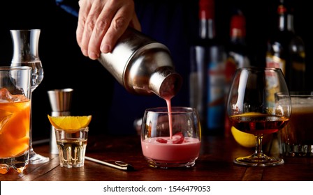 Bartender Pouring A Fruit Cocktail From A Shaker Into A Glass With Assorted Other Alcoholic Beverages On A Bar Counter In A Close Up View On His Hand In Shadowy Light