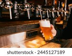 Bartender pouring light, cold foamy beer from tap into glass at bar with multiple beer dispensers. Serving craft beer from tap. Concept of alcohol drinks, nightlife, party, festivals, Oktoberfest. Ad