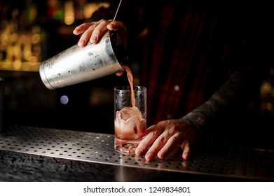 Bartender Pouring A Sippy Cup Cocktail From The Steel Shaker To A Glass With Big Ice Cube On The Bar Counter