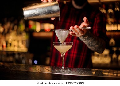 Bartender pourring a Daiquiri achoholic cocktail from the steel shaker through the sieve to a glass