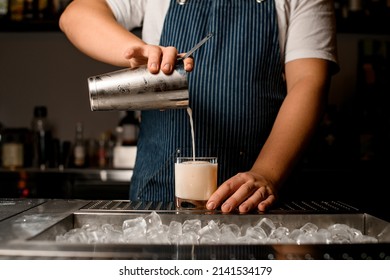 Bartender Pours Cocktail From Shaker Into Glass Standing On Metal Bar Counter. Shelves With Bottles Of Alcohol In Background.