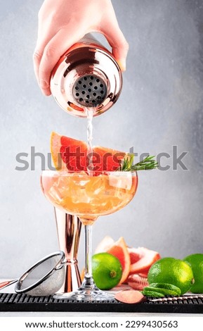 Bartender pours cocktail from shaker into glass. Grapefruit daiquiri alcoholic cocktail with white rum, syrup, fruit juice, lime and ice in garnished margarita glass. Gray background, bar tools