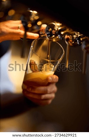 A bartender pours draft beer into a glass, foamy beer forming as the tap flows. Warm bar lighting and a soft blurred background create a cozy, inviting nightlife vibe.