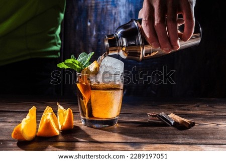 The bartender pours a drink from a shaker. Drink with ice and orange. Pour booze into a glass.