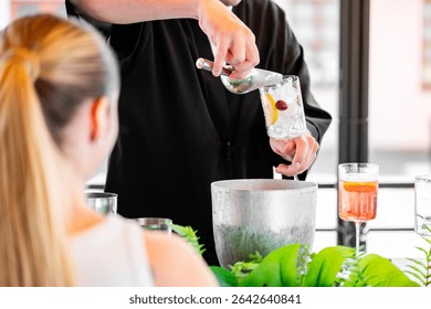A bartender prepares a cocktail, adding ice, lemon, and a cherry to a glass. A blonde woman sits nearby in a bright, modern bar setting with greenery.