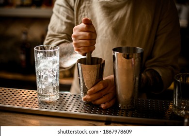bartender prepares cocktail in shaker cup using the muddler. Transparent glass with ice is next to it.