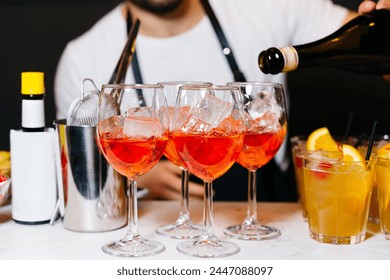 A bartender in the process of pouring a vibrant red aperitif into elegant wine glasses filled with ice, set against a bar backdrop.