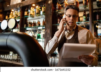 Bartender reading check list while talking on mobile phone and working in a pub.