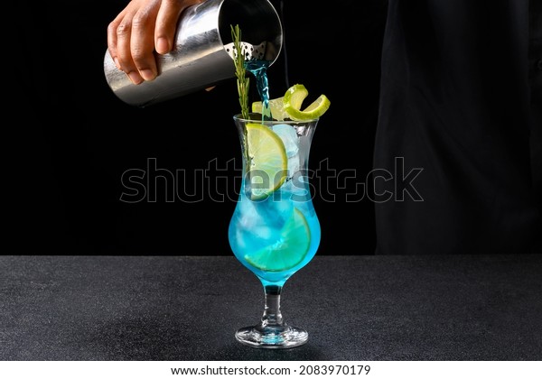 bartender serving with a cocktail shaker a\
blue drink in a glass decorated with rosemary, ice and lemon\
preparing a cocktail, on dark\
background.