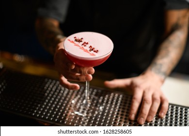 Bartender serving the pink alcoholic cocktail with a white scum decorated with the red pepper peas on the bar counter