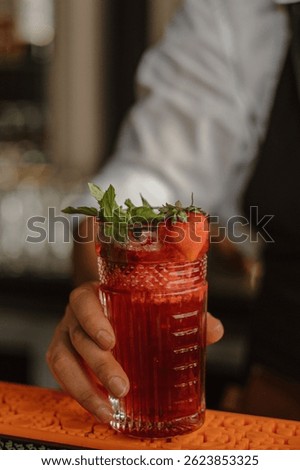 Bartender serving refreshing red cocktail with strawberry and mint garnish, close-up of hand holding summer drink at bar, fresh fruit beverage in crystal glass
