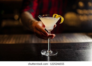 Bartender serving White Lady cocktail decorated with orange zest standing on the steel and wood bar counter on the blurred background