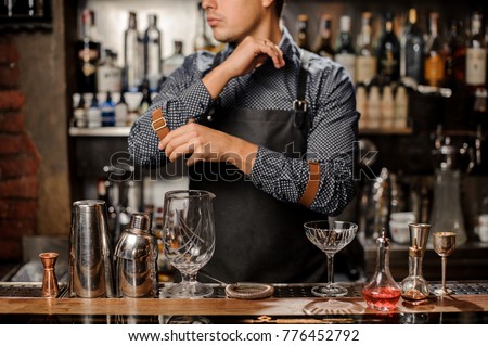 Bartender standing behind the bar counter with bar equipment and glasses on it and looking away