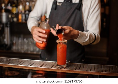 Bartender in the white shirt and dark apron pourring a bloody mary into the cocktail glass on the bar counter