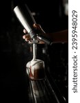 Bartender woman with steel whipping siphon accurate adds white foam to glass with cold coffee drink. Dark background. Selective focus