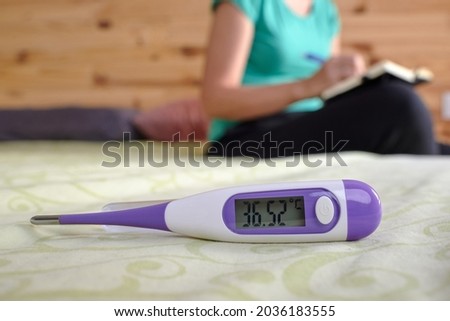 Basal thermometer with woman in background writing temperature