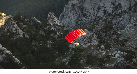 Base Jumper Under A Bright Red Parachute Flies Against A Background Of A Dark Rocky Landscape After Jumping From The Top Of A Mountain, View From Above. Panorama.