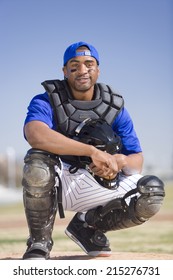 Baseball Catcher Wearing Blue Uniform, Cap, Protective Glove And Pads, Crouching On Pitch, Smiling, Front View, Portrait