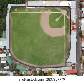 A baseball field sits surrounded by houses and streets in a neighborhood during the daytime.