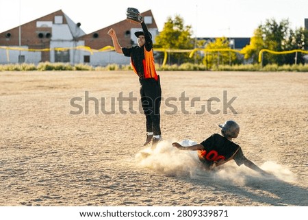 Baseball player sliding into base creating dust cloud while another player jumps to catch a ball, competing in a youth game