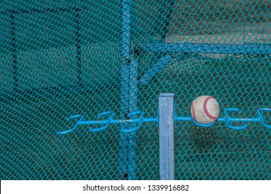 Baseball resting on metal bat rack in front of greenish blue netting around batter box. - Powered by Shutterstock