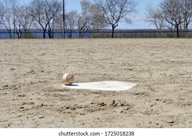 A Baseball Sits On Home Plate - With No People Around To Play The Sport.  Parks Have Been Closed To Public Gatherings And Both Professional And Amateur Sports Alike.