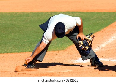 Baseball Umpire Dusting Off Dirt From Home Plate With A Brush With His Mask In His Other Hand