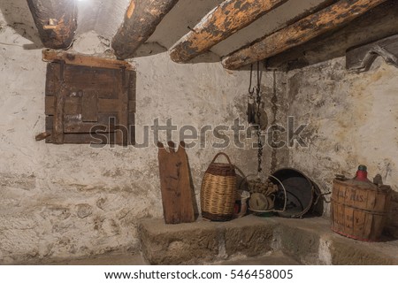 Basement of an old house with wooden beams and wall with humidity, antique household items, carboy demijohn, stewpot, plank, Wicker basket, pulley with chain, wooden window locked