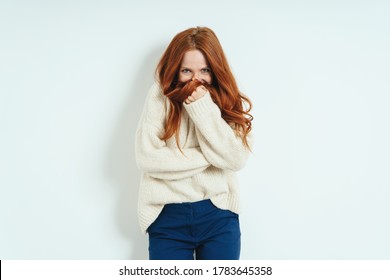 Bashful young woman hiding behind her hand and tress of her long red hair as she looks coyly at the camera against a white interior wall with copy space - Shutterstock ID 1783645358