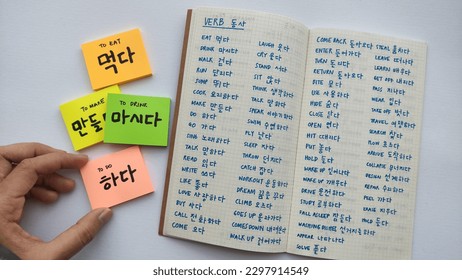 Basic korean word and verbs written in sticky notes and notebook. Simple words to remember for basic Korean language learning. White background, with a hand holding one of the sticky note.