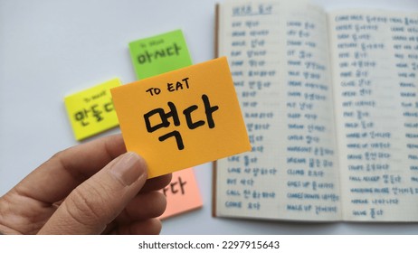 Basic Korean word and verbs written in sticky notes and notebook. Simple words to remember for basic Korean language learning. Focused on a hand holding a sticky notes with Korean verb.