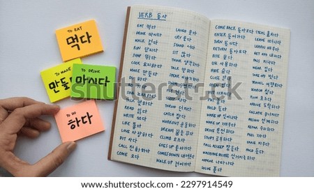 Basic korean word and verbs written in sticky notes and notebook. Simple words to remember for basic Korean language learning. White background, with a hand holding one of the sticky note.