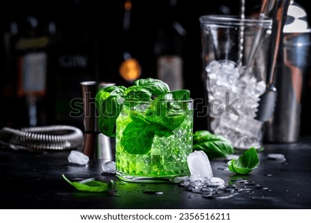 Basil smash green alcoholic cocktail drink with dry gin, sugar syrup, lemon, basil and ice, dark bar counter background
