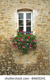 A basket of bright pink and red flowers hangs on the window of a home in an ancient building in France, surrounded by beautiful patterns in the stone wall.