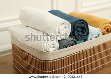 Basket with different rolled shirts on wooden table near white wall, closeup. Organizing clothes